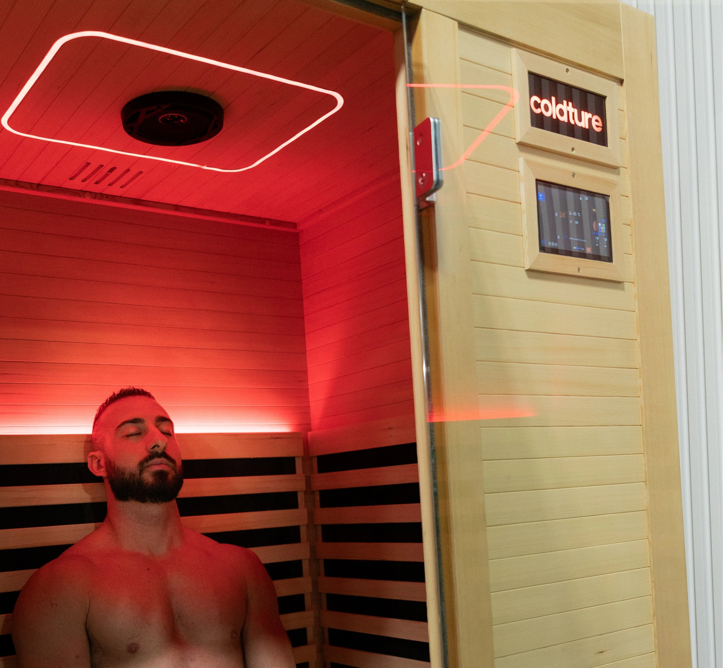 Man sitting inside a infrared sauna with red lighting, wearing dark shorts.