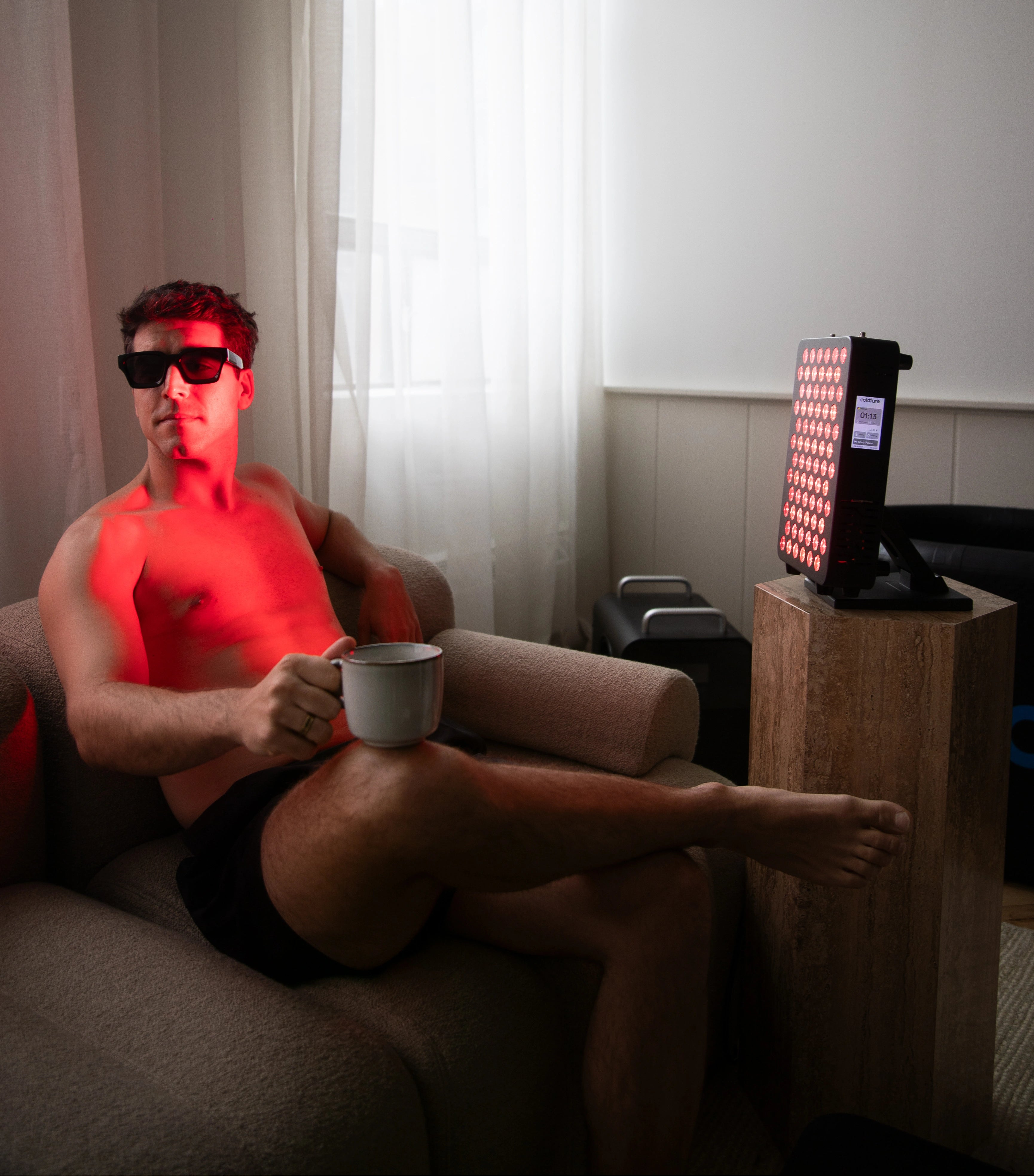 Person sitting on a couch with red lighting, holding a mug, in a room with a light curtain.